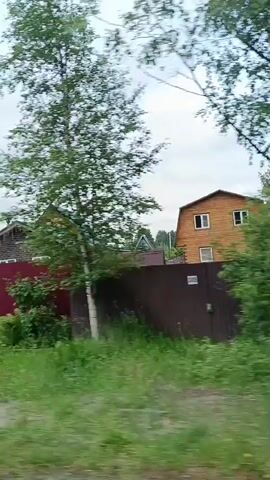 The video shows a view from a moving vehicle, likely a car, as it drives past houses and fences. The sky is cloudy, and there are trees and greenery visible. The houses appear to be in a rural or suburban setting. The first house visible has a wooden exterior and a prominent chimney. There is a large, dark red gate in the foreground of the initial frames. As the vehicle moves, different sections of fences and houses come into view. The fences are made of various materials, including corrugated metal and what appears to be wooden planks. Some parts of the fences are covered in greenery. The overall impression is of a drive through a residential area with a somewhat overcast sky.