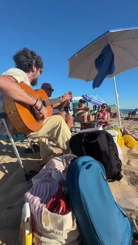 The video depicts a group of people on a sandy beach under a clear blue sky. In the foreground, a man with dark, curly hair and a beard is seated on a folding chair, playing an acoustic guitar. He is wearing a patterned shirt and light-colored pants. His left arm rests on the guitar, and his fingers are on the fretboard. To his right, another person, partially obscured, is also seated and appears to be clapping.

Further back, under beach umbrellas, two women are seated. One, wearing a bikini top, is also clapping. The other woman, wearing a pink top, is looking towards the guitarist. Several beach bags and blankets are visible in the foreground. The environment is clearly a beach with other beachgoers and umbrellas visible in the distance. The overall atmosphere appears relaxed and festive, suggesting a casual gathering with music.