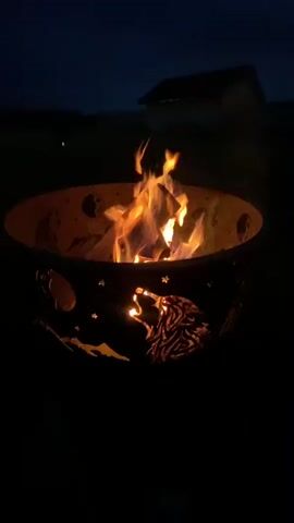 This is a close-up video of a fire pit at night. The fire pit has cutouts of a howling wolf, mountains, and stars. The flames from the fire are visible and are reaching upwards. The background is dark, indicating that it is nighttime, and there is a faint outline of a building on a hill in the distance. The video is shot from a low angle, focusing on the fire pit and the flames.