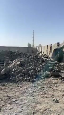 The video shows a field of rubble and debris under a clear, bright sky. In the background, there is a partial wall and a tall, thin minaret in the distance. As the camera pans slightly, more of the destroyed buildings come into view, revealing cracked and crumbling walls. One person dressed in white traditional clothing walks amongst the ruins. The ground is barren and dusty. The sun shines brightly.