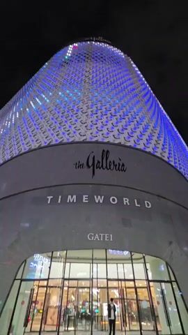 This low-angle, upward-facing video captures the exterior of The Galleria Timeworld in Daejeon, South Korea, at night. The building's distinctive, curved facade is illuminated by blue and white lights that create a dynamic pattern across its textured surface. The name "the Galleria" is prominently displayed in a stylish script on a grey band, with "TIMEWORLD" written in capital letters below it. Further down, "GATE 1" is visible above the arched entrance. The entrance itself is a large, glass-enclosed area with revolving doors, revealing interior lights and some blurry figures inside, including at least one person dressed in dark clothing standing near the entrance. The sky above is dark, indicating nighttime, and no specific weather conditions are apparent. The overall impression is one of a modern, well-lit commercial building at night, emphasizing its architectural design and prominent branding.