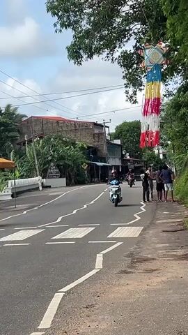 The scene unfolds on a road with multiple people either walking or riding motorcycles. A group of four individuals stand on the sidewalk to the right.  The road itself has a pedestrian crosswalk marked by thick white stripes.  On the left side of the road, two motorcycles can be seen riding away from the camera, while one motorcycle is closer, riding towards the camera. Another motorcycle can be seen further down the road also riding away from the camera.  Overhead, suspended from a tree, is a decorative hanging with a star-shaped top and colorful fringe, predominately white, red, and blue. The environment includes buildings, greenery, and a partly cloudy sky.  Power lines stretch across the sky in the background.