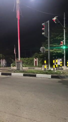 A night scene on a paved road with a traffic light, a lamppost, trees, and a sidewalk. A motorcycle with two riders passes by. There is also a white car moving in the distance.