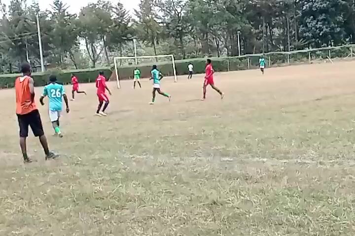 The video shows a soccer game in progress. The players are running on a grass field toward a goal. The players are wearing red and green jerseys. The field is bordered by trees and spectators.