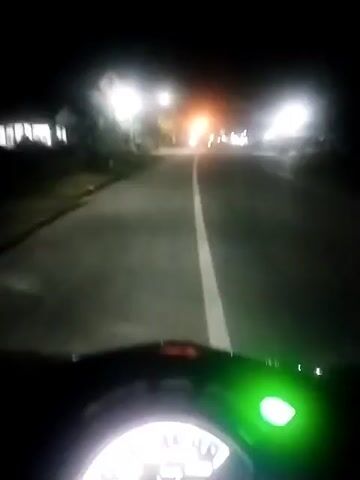 The video shows a night-time view from the perspective of someone riding a scooter or motorcycle on a paved road. The road has a single white line down the center. Streetlights illuminate the road, and there are houses visible on the sides. The dashboard of the vehicle is visible at the bottom of the frame, with a lit green indicator. In the distance, other vehicle lights can be seen. The camera is relatively steady.