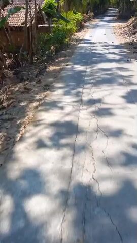 The video shows a concrete road in a rural area. The road is cracked in several places. There are trees and vegetation on both sides of the road, and a building is visible on the left. The sun is shining, casting shadows on the road. There are some fallen leaves on the road. In the distance, there appears to be a red and white striped arch or flag across the road. The camera is moving forward along the road.