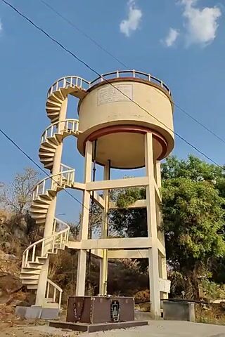 The video shows a water tower with a spiral staircase winding around it. At the base of the tower is a memorial stone decorated with garlands of flowers. The background consists of a rocky hillside, some trees, and a clear blue sky with wispy clouds. The video seems to be taken during the daytime, and the lighting appears natural. The focus is on the water tower and the memorial.