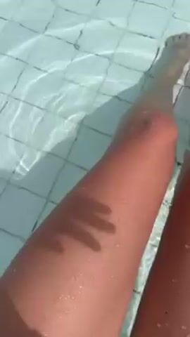 A first-person view shows two legs dangling in a swimming pool. The legs are tan and appear to be wet. The pool has a white tiled bottom, and the water is clear with slight ripples. The person touches their leg with their hand in the pool, creating more ripples. The scene is brightly lit, possibly by sunlight.