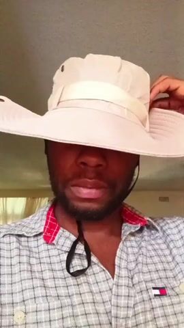A person is wearing a light-colored hat with a wide brim, pulled down low over their eyes. They are also wearing a gray and white plaid shirt with a red collar. The person adjusts the brim of the hat with their hand, revealing multiple bracelets on their wrist. The background appears to be an indoor space with light-colored walls and curtains.