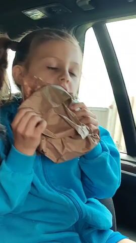 A young girl with blonde hair in pigtails, wearing a blue hooded jacket, sits in a car and eats from a brown paper bag. She is holding the bag with both hands and bites into something inside. The interior of the car is visible, with a window in the background showing a blurred outdoor scene.