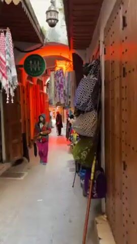 The video shows a narrow alleyway in a Moroccan market. The alley is lined with shops selling a variety of goods including textiles, bags, and baskets. The lighting is warm, with red and orange hues illuminating the scene. Several people are walking through the alley, including a person in a brightly colored jacket and pants. The shops are cluttered with merchandise, creating a vibrant and busy atmosphere.