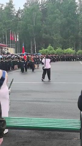 The video shows a parade with people in uniform marching, possibly for an official event or ceremony. The subjects are a group of individuals in dark uniforms with red accents, marching in formation. Some are carrying flags, including one with red and blue colors. They are on a paved area, likely a parade ground, with a line of trees in the background. Spectators are also visible, some holding umbrellas, suggesting it might be raining or drizzling. The lighting indicates it is daytime, possibly late morning or afternoon. The atmosphere appears formal and orderly, typical of a military or educational institution's parade. There is no visible text in the video. The scene is set in an outdoor environment, with a building and flags visible in the distance, possibly indicating a school or military base in Parechcha, Belarus.