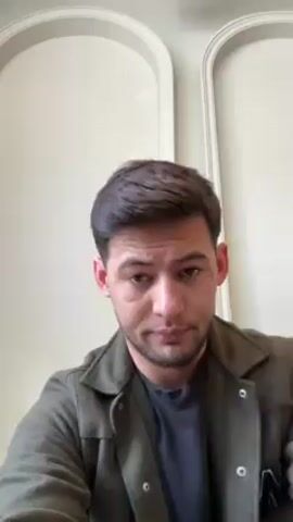 A man faces the camera directly. He wears a green collared jacket over a dark-colored shirt. He touches his chin briefly and then runs his hand through his hair. The background consists of a plain wall with two framed, arched designs.