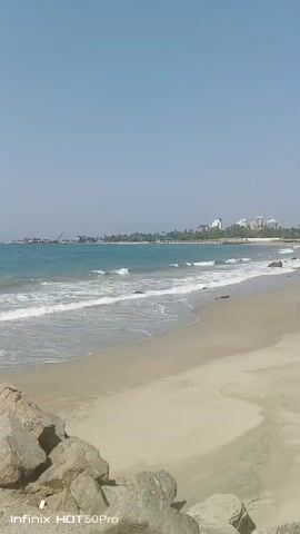 The video shows a sunny day at the beach. There are waves rolling onto the sandy shore. In the background, there are several buildings, some trees, and what appears to be a pier. In the lower left corner, there are some large rocks. The sky is clear and blue.