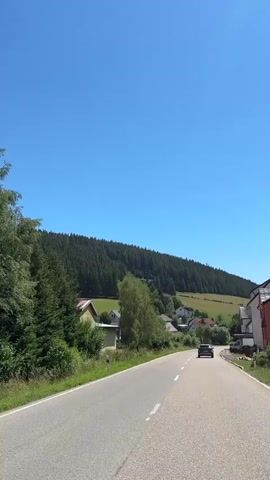 A car drives down a paved road with a line of trees and houses on the right. A hill covered in trees is visible in the background under a clear blue sky.