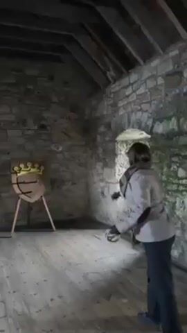 A woman throws metal rings at a target in an enclosed room, likely a castle or stone structure. The woman has short brown hair and wears a gray coat, black pants, and dark shoes. She stands on a wooden floor with a light gray finish. The room is small and contains stone walls, a window with an arched top, and a wooden ceiling with exposed rafters. The target is a shield-shaped object with a crown design on it, mounted on a wooden stand.  The rings are thrown underhand.  One throw results in a clang as it hits the target.