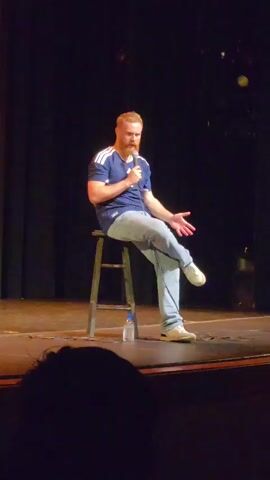 A man with red hair and a beard, wearing a dark blue t-shirt with white stripes on the sleeves and light blue jeans, sits on a dark stool on a stage. He is holding a microphone in his right hand and gesturing with his left hand. A bottle of water is on the floor to his left. The background is a dark curtain. The stage floor is visible in the foreground, and the silhouette of an audience member's head is in the lower part of the frame. The lighting appears to be from stage lights, illuminating the performer and the immediate area around him.