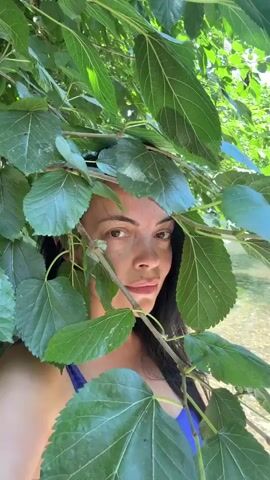 A medium shot shows a woman with dark hair and brown eyes peeking through the leaves of a tree. She is wearing a blue bikini top and looking directly at the camera with a slight smile. The background shows a body of water with rocks and sunlight reflecting on the surface. The leaves are large and green, obscuring parts of her face and body.
