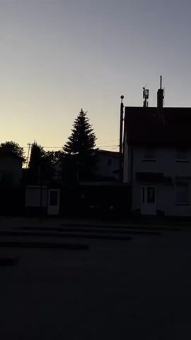 The camera pans right across a quiet street towards a white two-story building with a red roof under a dim sky. Two figures can be seen sitting on a bench in front of the building. As the camera continues to pan right, a motorcycle comes into view. The scene appears to be a small town square or parking lot at dusk.