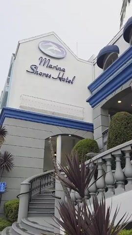 The video shows the exterior of the Marina Shores Hotel. The building has a white facade with blue trim and awnings over the windows. A sign on the building reads "Marina Shores Hotel" with a logo of a stylized bird. There is a grand staircase with a balustrade leading up to the entrance. Lush green bushes and palm trees are visible around the entrance. The sky is overcast, suggesting a cloudy day. The camera pans upwards, capturing different angles of the hotel's architecture and surrounding greenery. No people are visible in the video.