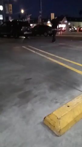 The video depicts an outdoor scene at night. The main focus is a parking lot with double yellow lines. A yellow parking stop barrier is prominently positioned in the foreground. Several cars are parked in the background, with street lights illuminating the area. A person is walking between the cars. The overall atmosphere appears quiet. The video is a static shot with no camera movement.