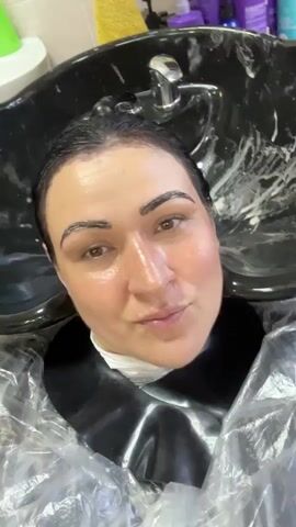 A close-up shot of a woman's face as she lies in a salon hair washing station. She is wearing a black cape and a clear plastic covering around her neck. Her eyebrows are dark and appear to have product applied to them. White foam is visible in the sink behind her head. She has dark hair that is wet. The lighting suggests an indoor environment, likely a hair salon.