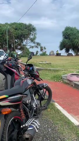 A close-up shot of a parked motorcycle is in the foreground. Behind the motorcycle, a grassy field extends to the ocean. A person is sitting on a bench in the distance, facing the ocean. The sky is cloudy.