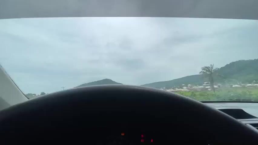This video captures a forward-facing view from inside a vehicle, likely a car, as it travels along a paved road. The scene is a rural or semi-rural environment with a small village on either side of the road, nestled at the base of rolling, tree-covered hills. The architecture of the village appears to consist of various houses with distinct roofs, some conical and others more rectangular.

The road itself is a two-lane asphalt surface, with a reddish-brown dirt shoulder. In the distance, the road curves gently and is lined with more village structures and greenery. Several vehicles are visible on the road, including a large white tanker truck with blue and yellow stripes passing the viewer's vehicle on the left. Further ahead, a white truck with a blue tarp is traveling in the same direction. There are also a few people visible walking or standing near the roadside.

The sky is overcast with a layer of grey clouds, suggesting a cloudy day. The overall lighting is diffused and even, indicating it is likely daytime, possibly late morning or early afternoon. The presence of lush green vegetation indicates a potentially tropical or subtropical climate. Power lines with poles are visible on the left side of the road, hinting at some level of infrastructure.

The mood of the scene is calm and observational, typical of a travelogue or documentary footage. There is no discernible text within the video frame. The setting appears to be Koton Karifi, Nigeria, as indicated by the prompt.