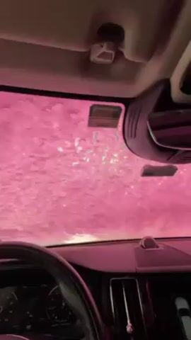 The view is from inside a car going through a carwash. Water and soap cover the windshield. The soap changes color, transitioning between pink, blue, and green. The steering wheel and dashboard of the car are visible.