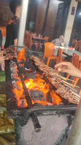 The video shows a close-up of satay skewers being grilled over hot coals. Flames are visible, and the meat appears to be well-cooked and slightly charred. In the background, people are seen sitting at tables, suggesting a restaurant or outdoor food stall setting. The lighting is dim, indicating it is likely evening or nighttime.