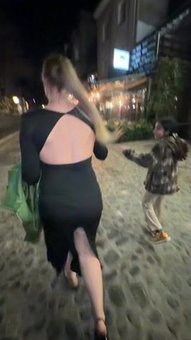 The video shows three people walking down a cobblestone street at night.  A woman in a black dress with an open back walks in the foreground, followed by a young girl in a camouflage jacket and beige pants who reaches out to touch her. A woman holding a bouquet of red roses looks into the camera and smiles. The street is lined with restaurants with outdoor seating areas lit by strings of lights.  A building with flags is seen in the background. The video appears to be shaky and taken with a mobile phone.