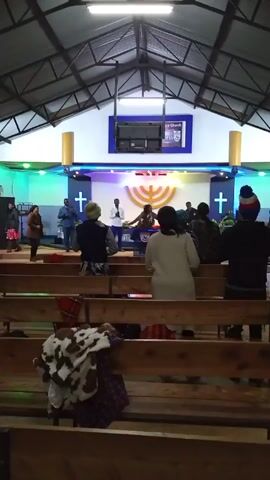 A church congregation is standing and singing during a service. The church has wooden pews and a stage with a cross and a menorah. There are approximately a dozen people in the video. Some people are standing and facing the stage, while others are sitting in the pews.