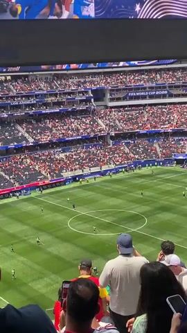 The video shows a wide view of a soccer stadium packed with spectators. In the foreground, the backs of several people are visible, looking towards the soccer field. The field itself is a vibrant green with white markings, and several players in dark uniforms are scattered across it. Above the field, a large Jumbotron screen displays a group of cheerleaders in blue and yellow uniforms, dancing energetically. The screen also shows the FIFA World Cup 2026 logo. The stadium seating is filled with fans, many wearing red. The overall atmosphere appears to be festive and anticipatory, suggesting a significant sporting event is about to take place or is underway.