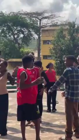 A group of about a dozen young people is gathered outdoors on a paved surface, possibly a school courtyard or park, with trees and a multi-story building visible in the background. The people are diverse in appearance and are dressed in casual clothing, including t-shirts, tank tops, jeans, and leggings. They appear to be conversing and gesturing with their hands, suggesting they are engaged in a discussion or activity. The overall atmosphere seems relaxed and informal. The weather appears to be sunny and pleasant.