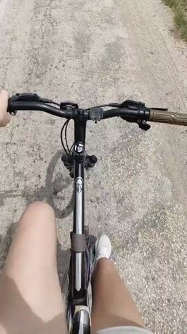 The video shows a first-person perspective of someone riding a bicycle on a dirt path. The rider's legs and hands are visible, as well as the handlebars and front of the bicycle. The path is surrounded by dry grass and vegetation, with trees and a field in the background. The sky is blue with white, fluffy clouds, suggesting it is a sunny day. The overall mood appears to be one of peaceful enjoyment of an outdoor activity. The video seems to be taken in the vicinity of Ароматновское сельское поселение, Ukraine. No text is visible in the video.