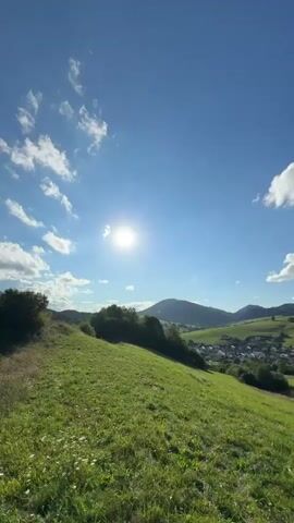 This is a vertical, wide shot of a vast landscape under a bright, sunny sky. The sun is positioned high in the sky, slightly off-center to the right, casting a warm glow. Wispy white clouds are scattered across the clear blue sky. The foreground is dominated by a gently sloping grassy hill covered in green grass and some taller, dried grasses and wildflowers. A dark green, bushy shrub is visible on the left side of the hill. In the middle ground, the hill continues to slope downwards, revealing more green vegetation and a line of trees. Further in the background, rolling hills covered in a mix of green trees and fields form a picturesque valley. A small village with scattered houses and buildings can be seen nestled within the valley, with roofs of various colors. The scene evokes a sense of peace and natural beauty.