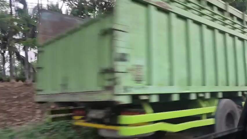 A green truck drives past. A man in a black shirt and a person in a black shirt walks in front of the truck. Trees and buildings are visible in the background.