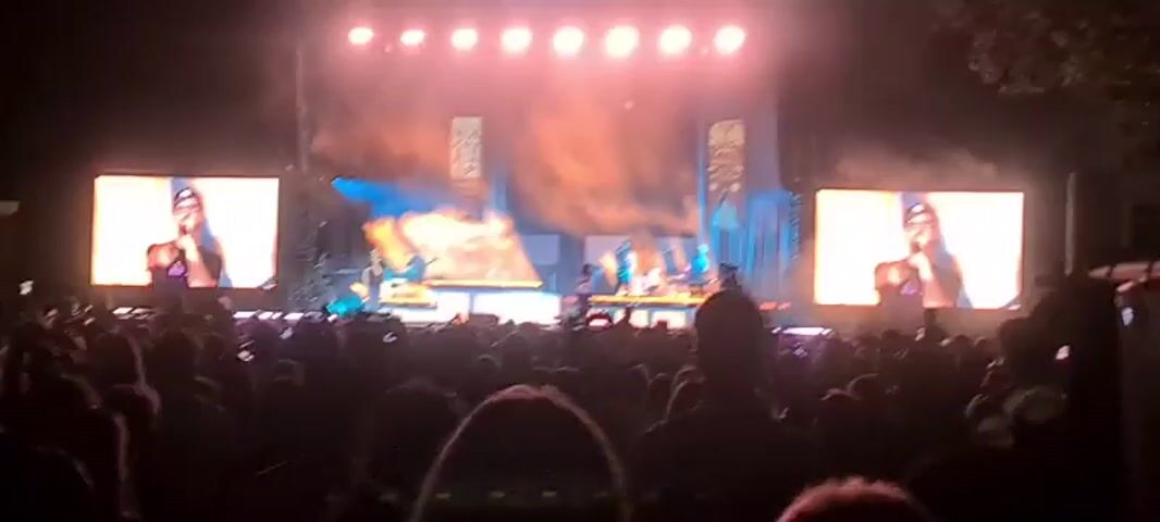 The video shows a live concert performance. A band is on stage with multiple musicians playing instruments and singing. There are large screens on either side of the stage displaying close-ups of the performers. A large audience is gathered in front of the stage, many holding up their phones to record the event. The stage is lit with bright spotlights and there is fog or smoke effects. The overall atmosphere is energetic and festive.