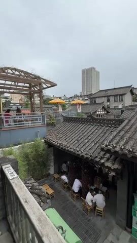 The video presents a high-angle, panoramic shot of a traditional East Asian architectural complex with dark tiled roofs and brick walls, under an overcast sky. The camera pans slowly from left to right. In the foreground, several individuals are seated at small wooden tables and chairs in an outdoor area. A wooden pergola with climbing plants is visible on the left. Two large yellow parasol-like structures are situated on a distant roof. A stack of green and white Starbucks boxes is visible on the right. As the camera pans, an archway in a brick wall and additional traditional buildings come into view. Beyond the rooftops, vibrant pink flowering trees are prominent. A tall, light-colored modern skyscraper is visible in the far background.