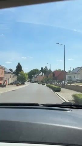 The video shows a view from inside a vehicle traveling down a paved road through a suburban or semi-rural area. The road is clear of traffic. On the right side of the road, there are several houses with pitched roofs, manicured hedges, and trees. A street light is visible on the right side. On the left side of the road, there are more houses, some trees, and a parked car. The sky is clear and blue. The camera is angled slightly downwards towards the dashboard of the vehicle. The video is shot during daylight.