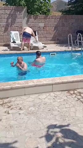 At 00:00, a person wearing American flag shorts stands by a swimming pool. At 00:01, another person in the pool splashes water. At 00:02, the person in the flag shorts turns their back to the camera. At 00:03, one person jumps into the pool. At 00:04, a person sits on the edge of the pool. At 00:05, a person in the pool gestures with their hands. The scene is a swimming pool with a brick wall and lounge chairs in the background.