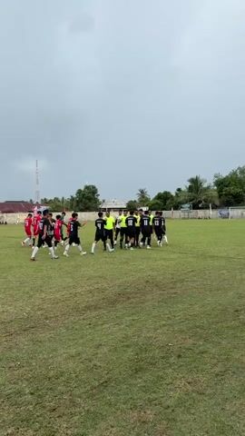 This video shows a group of male soccer players on a grassy field. They are gathered in the center of the field, some wearing black and white uniforms, others in red and white, and a few in neon yellow. One player in the black and white uniform is holding a soccer ball. Some players appear to be shaking hands or interacting with each other. The sky is overcast, and there are trees and buildings in the background. The grass appears somewhat worn.