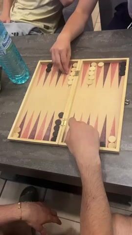 A group of people are playing backgammon at a table. The camera is positioned above the table, looking down at the game board. The backgammon board is made of wood and has the standard triangular markings. There are black and white pieces on the board, arranged according to the rules of the game. Two people's hands are visible, one moving a black piece and the other reaching for a die. A blue water bottle is on the left side of the table. The environment appears to be indoors, possibly a home or a casual dining establishment.