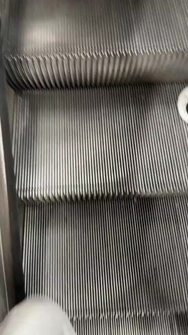 This video captures a person ascending an escalator in a public transportation setting, likely a subway or metro station in Şişli, Turkey. The escalator steps are made of metal and are moving upwards. The person in focus is wearing a long white dress with a subtle pattern and a dark green hooded cloak draped over their shoulders. The cloak has a decorative white and silver band across the chest.

The background reveals a bustling station environment. Other people are visible on the escalator and in the surrounding area, suggesting it is a busy time. The station architecture appears modern, with a metallic ceiling and overhead lighting. The lighting is bright and even, typical of an indoor, underground space.

The activity is simply people using the escalator to travel. The video focuses on the movement and the perspective of ascending. There are no strong emotional cues conveyed; the overall impression is one of routine transit. The time of day and weather are not discernible from the video content, as it is an indoor scene. There is no visible text within the frames. The context of Şişli, Turkey, is provided as an external detail, not directly evident from the visuals themselves.