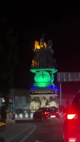 A nighttime shot captures a towering monument in the center of a bustling traffic circle. The monument is intricately carved, featuring multiple levels of sculptures, including elephants at the base, followed by a peacock design, and topped with various animal figures. The monument is illuminated with the colors of the Indian flag: orange at the top, green in the middle, and blue at the bottom. Several cars are visible in the foreground, with their headlights and taillights glowing in the dark. A person on a motorcycle drives past the monument on the left. The scene is well-lit by artificial lights, highlighting the monument and the surrounding traffic. The sky above is dark, indicating that it is nighttime.