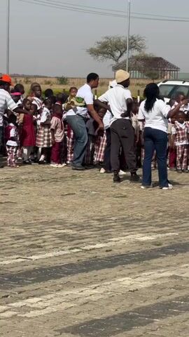 The video shows a group of young students, mostly in maroon and plaid uniforms, gathered in what appears to be a paved parking lot.  Adults are present, possibly teachers or chaperones, who seem to be organizing or directing the children. Several cars are parked nearby.  The sky is clear and the weather seems sunny.  The environment looks like it could be a school or event venue, with streetlights visible in the background. The children are standing, waiting, and interacting with each other, while the adults are moving around, potentially giving instructions or managing the group.