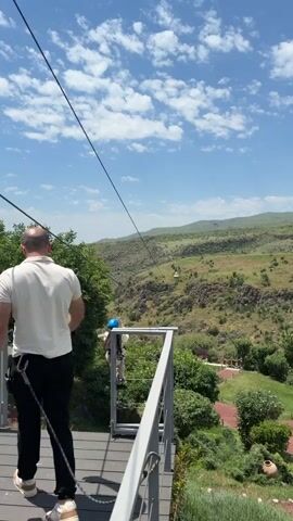 The video captures a person ziplining across a scenic valley. The view begins from behind a man standing on a wooden platform, looking towards a zipline. The camera then pans to follow a person ziplining over a steep, green valley with rocky outcrops. The zipliner, wearing a blue helmet, is suspended mid-air, appearing to enjoy the ride. The background shows a vast expanse of rolling hills under a bright blue sky dotted with white clouds. The zipline continues across the valley towards a distant platform. Lush green trees and vegetation cover the slopes. The overall scene conveys a sense of adventure and natural beauty.