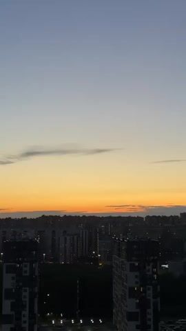 The video shows a time-lapse of a sunset over a city. The sky transitions from a pale blue to orange and then pink hues as the sun dips below the horizon. Silhouettes of numerous apartment buildings are visible in the foreground, with lights beginning to illuminate in some of the windows. A few wispy clouds drift across the sky. The city appears densely populated with many tall buildings. The perspective is from a high vantage point, looking down on the cityscape.