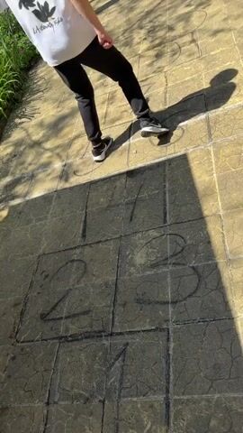 A person is playing hopscotch on a paved surface with painted numbers in squares and circles. The person is wearing black pants, black and white shoes, and a white t-shirt. They hop and reach down to touch the number seven written in a circle. The surface is made of square stone tiles and is partially in sunlight and partially in shadow. Grass and trees are visible in the background. There are no audible sounds in the video.