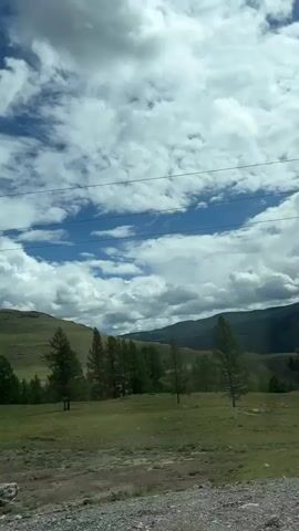 The video shows a rural landscape with rolling hills and scattered trees. The sky is partly cloudy with patches of blue visible. Power lines are strung across the top of the frame. The ground in the foreground is a mix of dirt and gravel, with a few large rocks. There are no people visible in the video. The camera appears to be moving, suggesting it is being filmed from a vehicle.